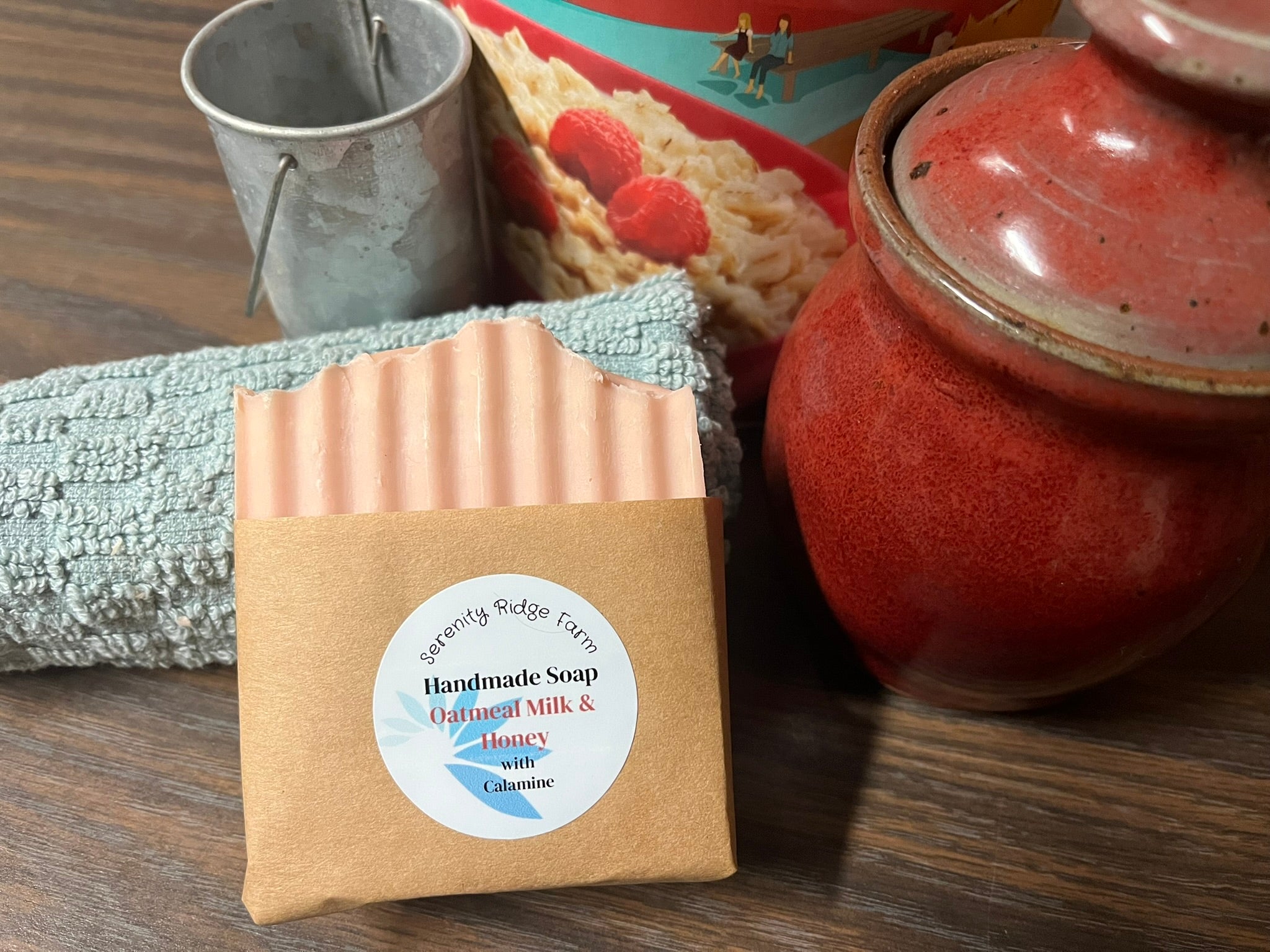 Handmade soap bar with a label on a wooden surface next to a red ceramic jar and a metal pail.