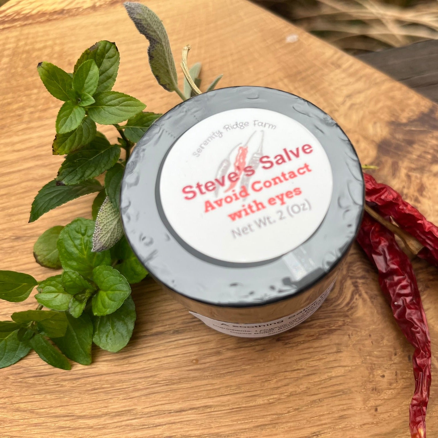 Jar of salve with a label on a wooden surface with plants and chili peppers.